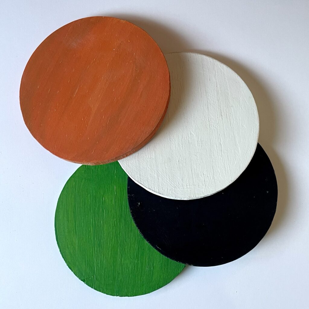 Four wooden coasters painted a base color of: orange, white, black, and green.